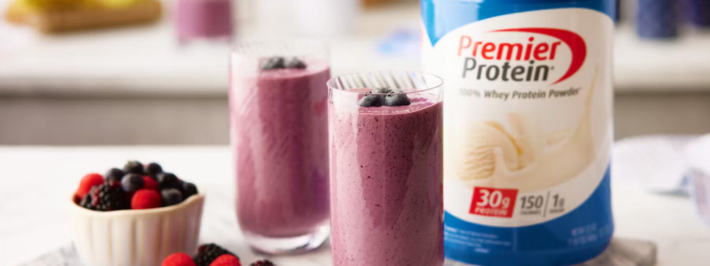 Two berry protein shakes in glasses sitting next to a tub of Premier Protein Vanilla Milkshake powder.